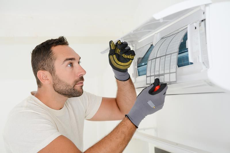 Un technicien installe un climatiseur mural à l'aide d'un outil, portant des gants, dans une pièce lumineuse.