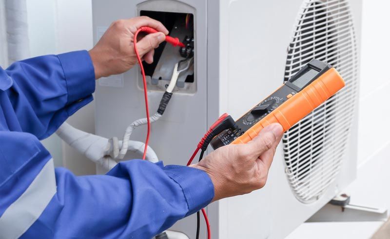 Un technicien teste un climatiseur extérieur à l'aide d'un multimètre et d'une pince ampèremétrique.