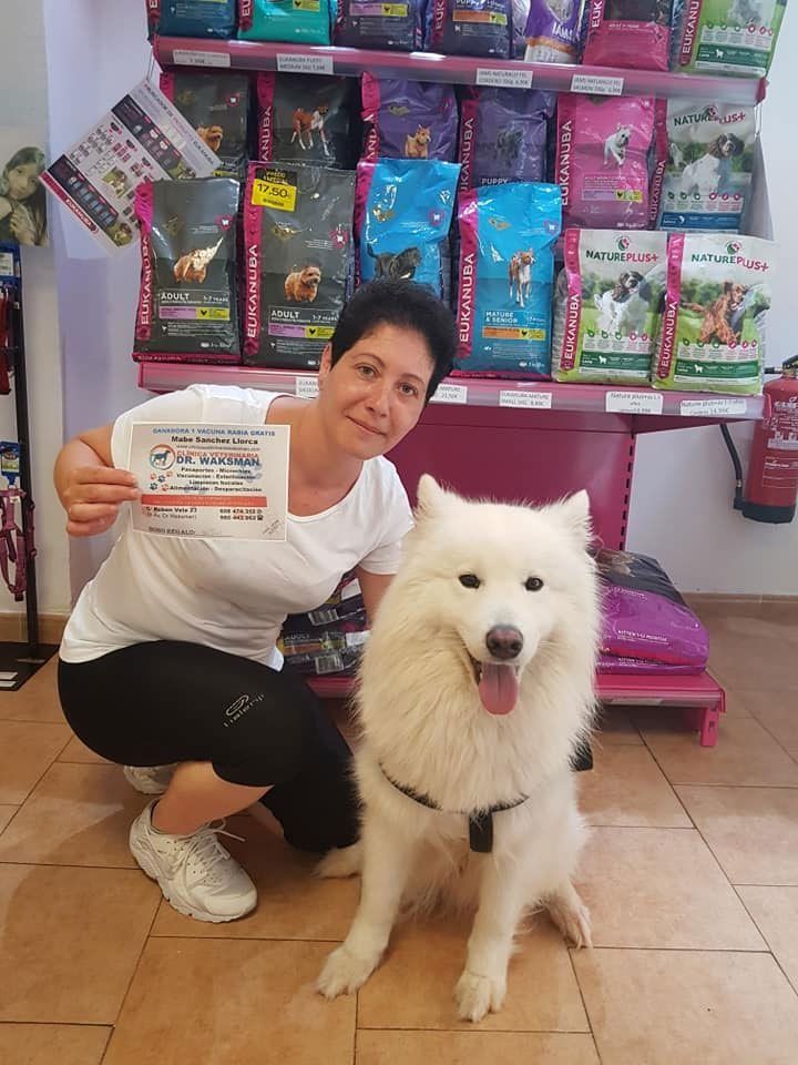 Mujer y perro blanco y esponjoso en una tienda de mascotas, con un certificado en la mano. Estantes de comida para mascotas detrás.