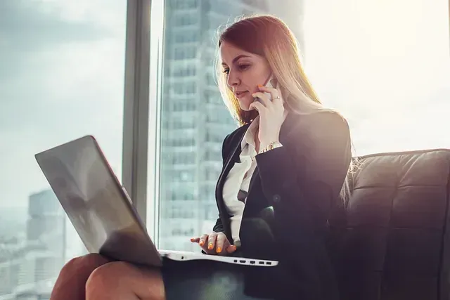 Una mujer está sentada en un sofá usando una computadora portátil y hablando por un teléfono celular.