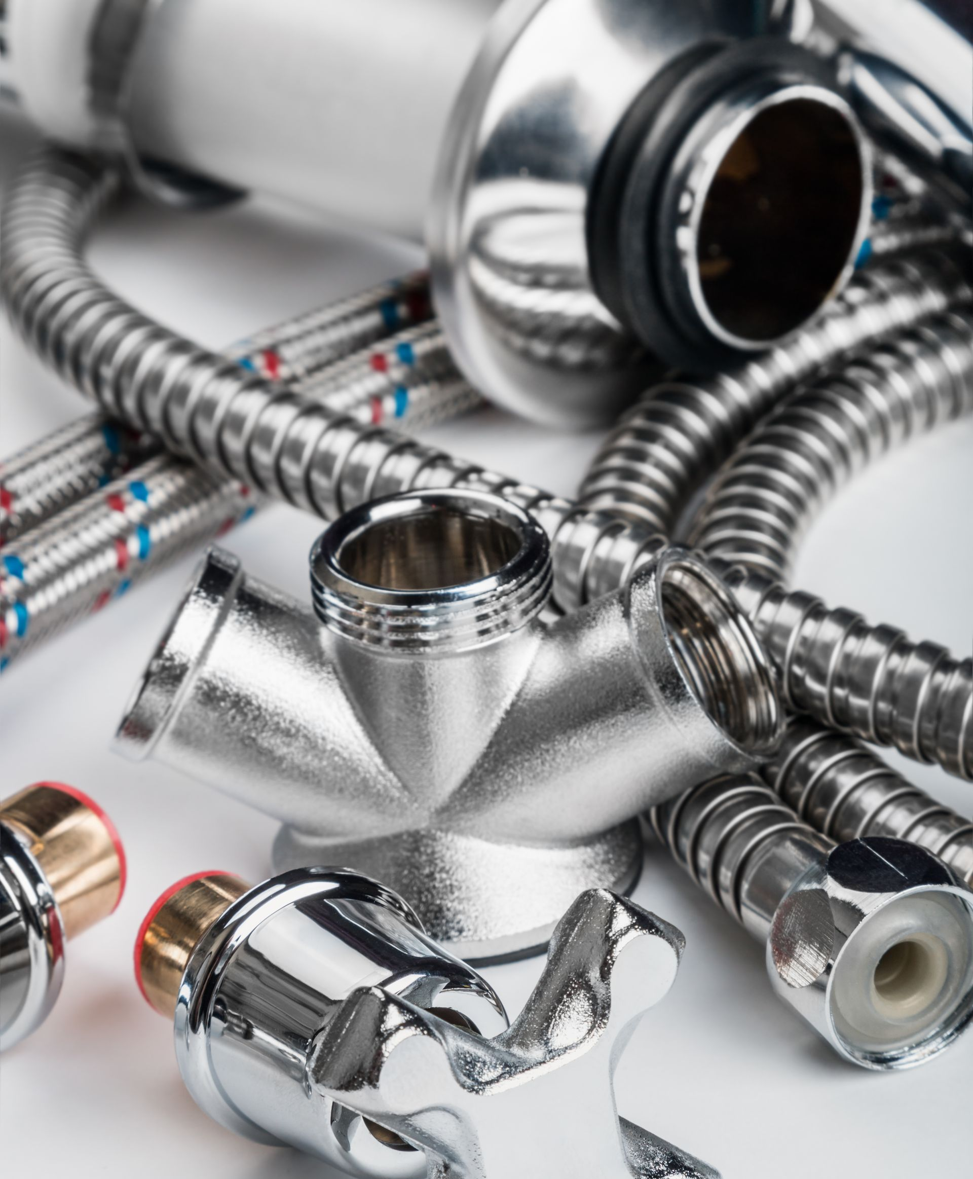 Accessoires de plomberie chromés, comprenant un raccord en T, des tuyaux tressés et des pièces de robinetterie, disposés sur une surface blanche.