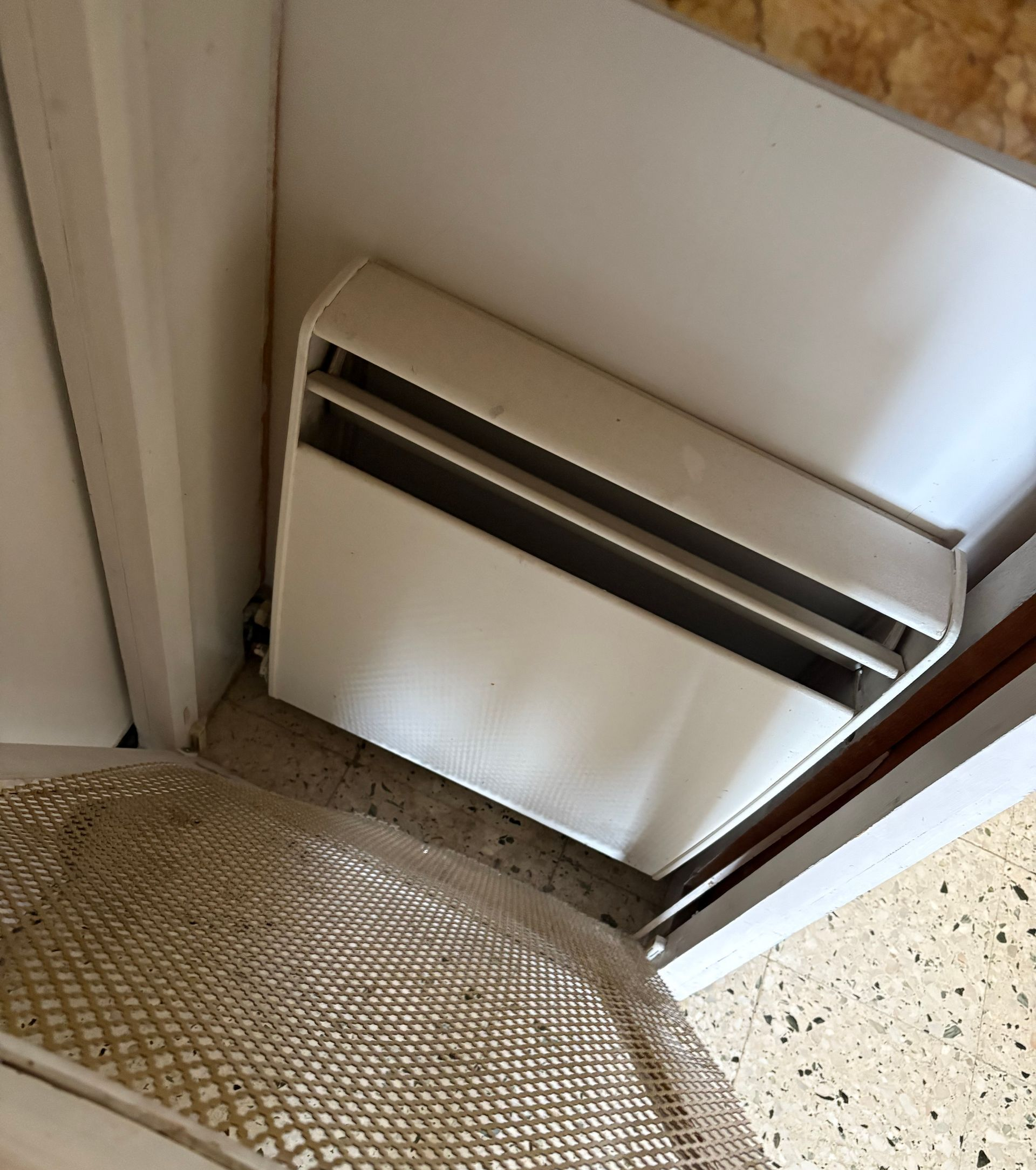 Un radiateur électrique blanc de type plinthe est placé dans un coin, à côté d'une chaise en osier, sur un sol carrelé.