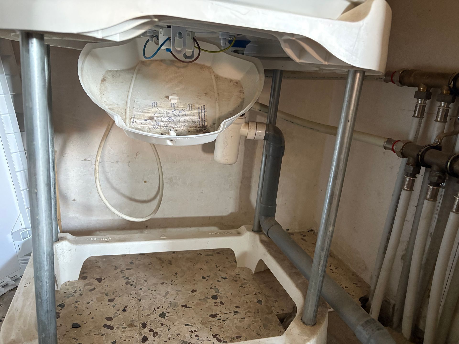 Évier de buanderie blanc, structure métallique, tuyauterie apparente. L'évier est taché de saleté.