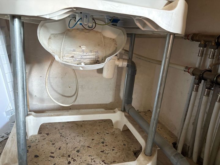 Évier de buanderie blanc, structure métallique, tuyauterie apparente. L'évier est taché de saleté.
