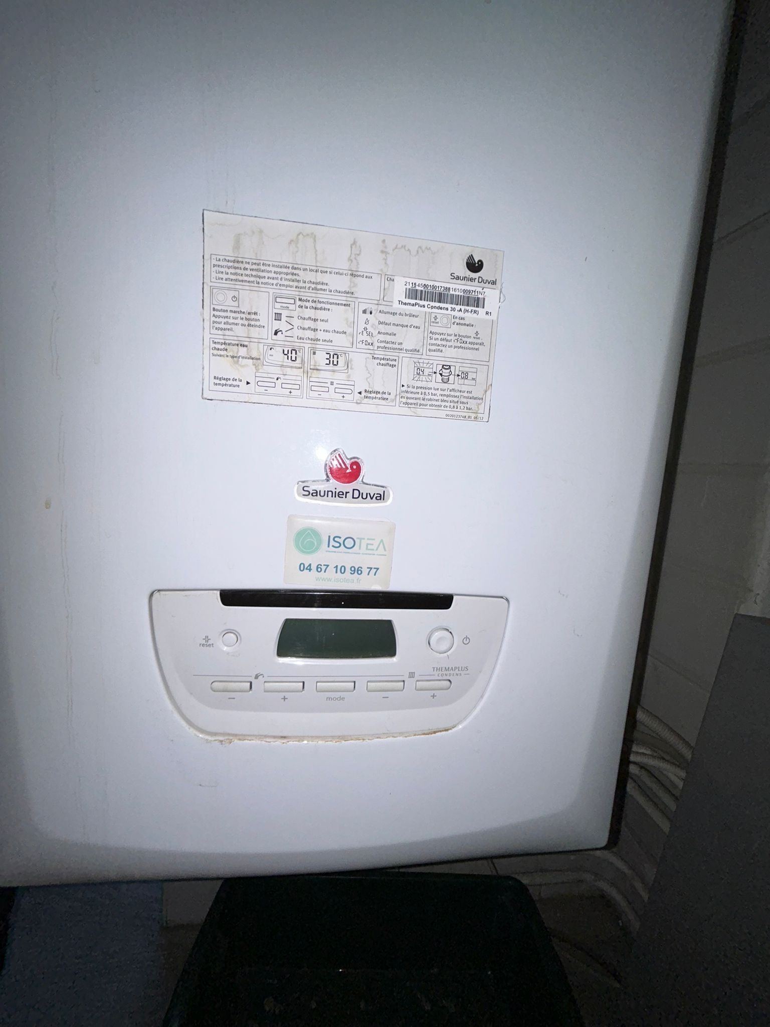 Chaudière à gaz murale blanche avec panneau de commande et écran, divers autocollants sur la face avant.