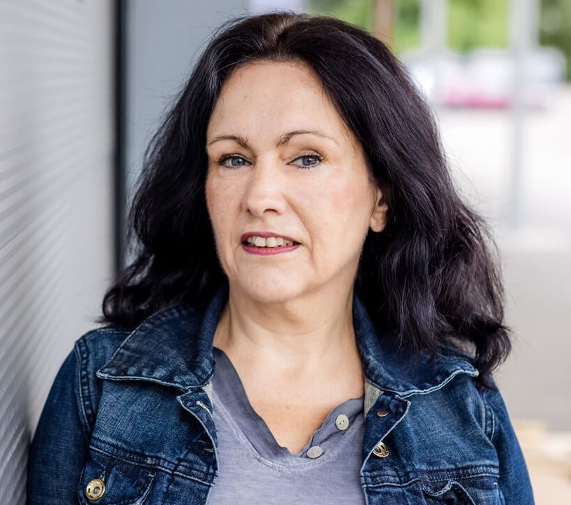 Frau in Jeansjacke, blickt in die Kamera, dunkles Haar, neutraler Gesichtsausdruck, Außenaufnahme.