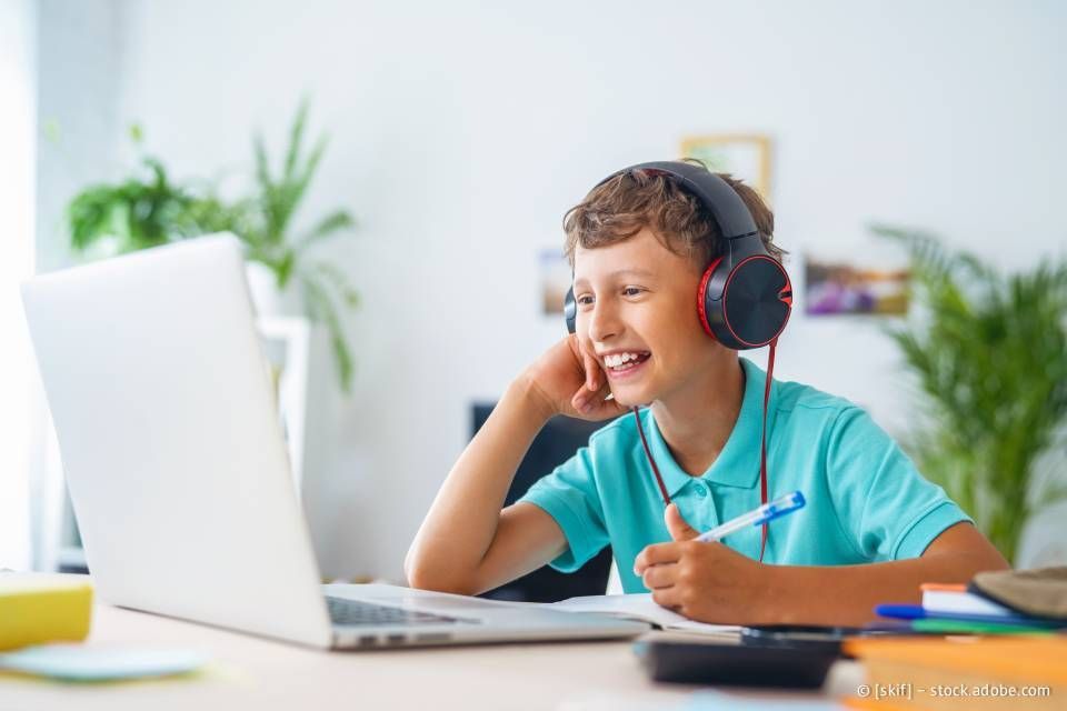 Ein Junge mit Kopfhörern lächelt auf einen Laptop und zeigt den Daumen nach oben. Drinnen, Schreibtisch mit Büchern und Stift.
