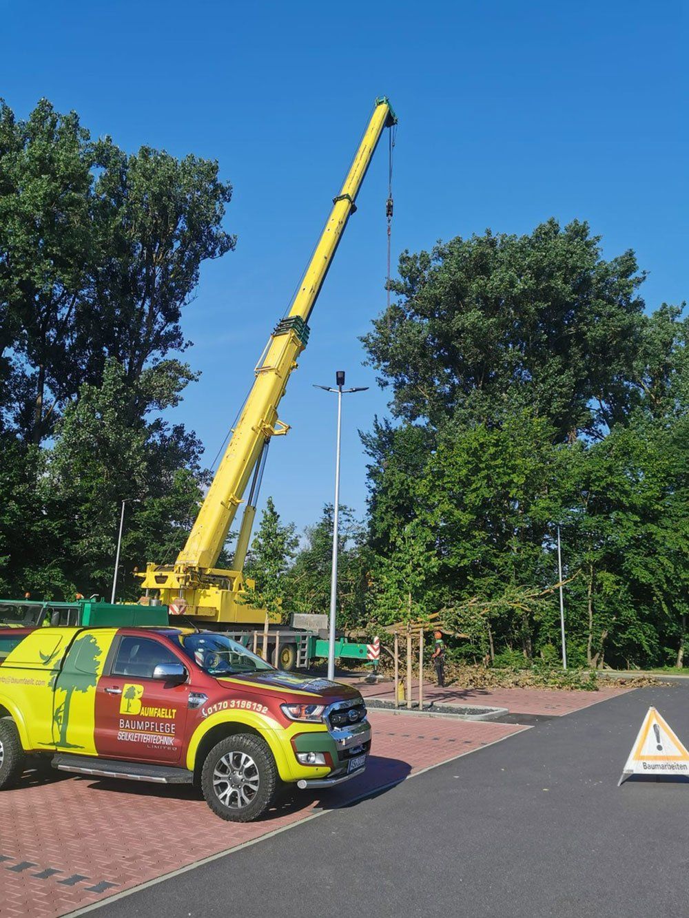 Neben einem Spielplatz steht ein gelber Kranwagen mit ausgefahrenem Ausleger. Blauer Himmel und Bäume bilden den Hintergrund.