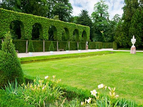 Gepflegter Garten mit leuchtend grünem Rasen, geschnittenen Hecken, bogenförmigen Laubstrukturen und einer dekorativen Vase.