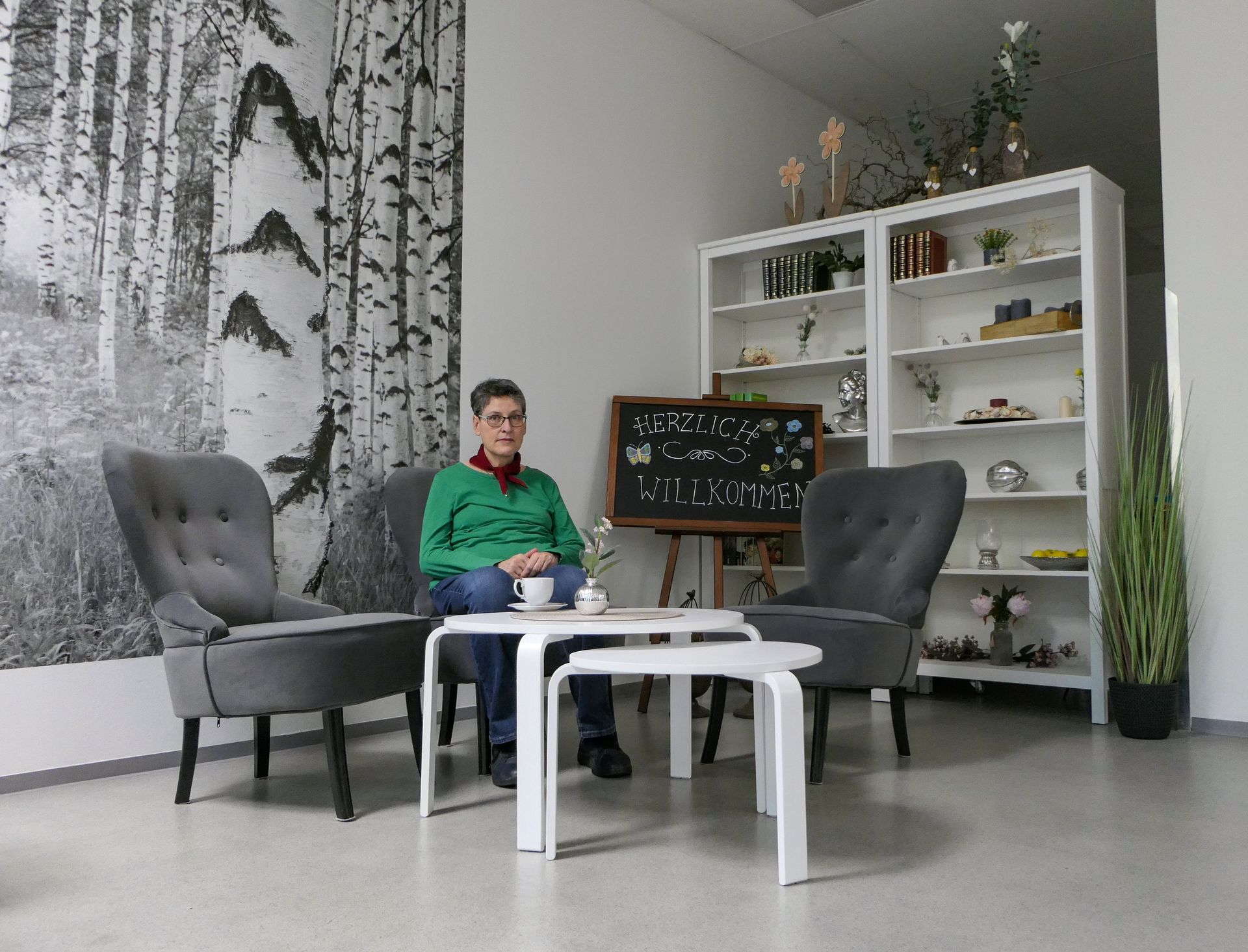 Sitzecke mit drei Sesseln, auf einem eine Frau, im Hintergrund eine Tafel mit Aufschrift: Herzlich Willkommen