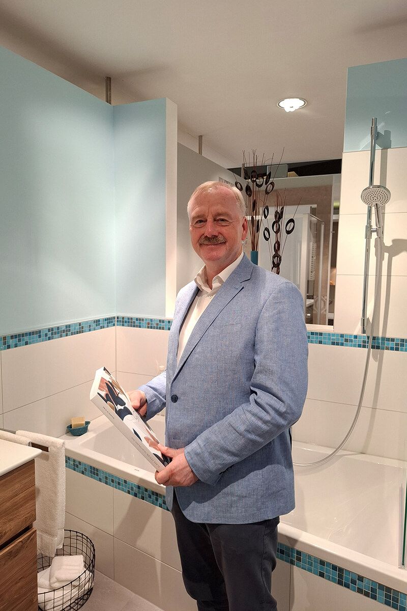 Mann im Blazer hält eine Zeitschrift in der Hand und steht in einem Badezimmer mit blauen Akzenten und Badewanne.