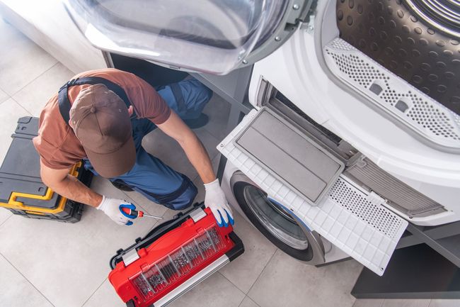 Un technicien nettoie une machine à laver avec un aspirateur rouge à côté du tambour ouvert et du bac à lessive.
