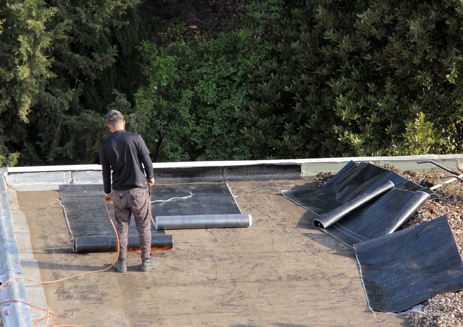 Un ouvrier installe une membrane d'étanchéité noire sur un toit plat entouré d'arbres verts.