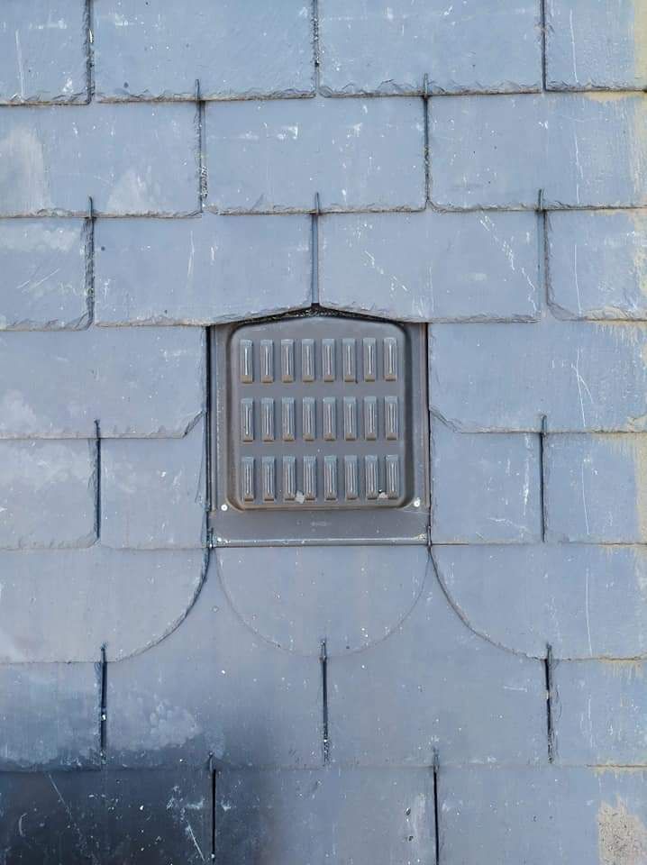 Gros plan sur le toit d'une maison avec des tuiles couleur terre cuite, une cheminée en pierre et une lucarne cintrée, le tout sur fond de ciel bleu.