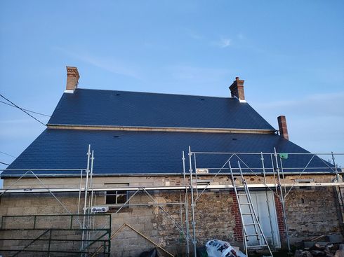 Gros plan sur le toit d'une maison avec des tuiles en terre cuite rouge, deux lucarnes et une cheminée en briques sur fond de ciel bleu.