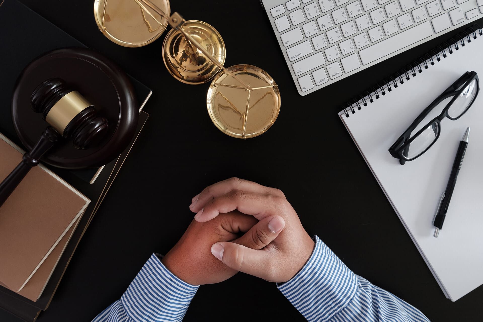 Escritorio con manos entrelazadas, relojes dorados, auriculares, cuaderno, gafas y teclado sobre un fondo negro.