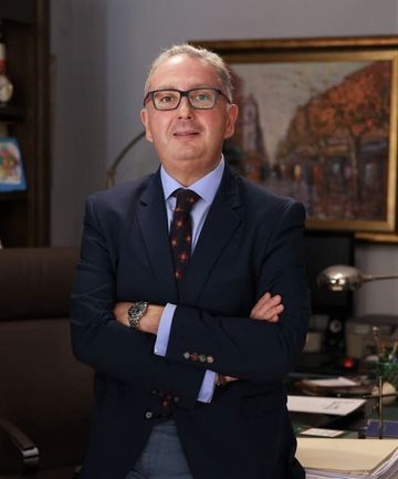 Un hombre con traje y corbata está de pie frente a un escritorio con los brazos cruzados.