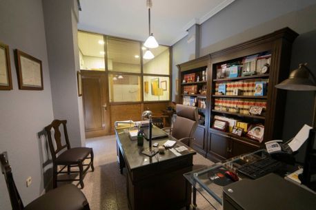 Despacho en casa con escritorio, silla, estantería y mampara de cristal en una habitación con iluminación cálida.