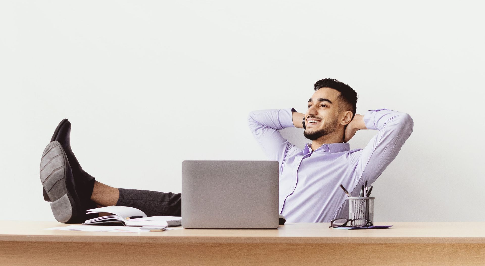 Mann sitzt am Schreibtisch, verschränkt die Arme hinter dem Kopf, legt die Beine auf den Tisch und lächelt erleichtert