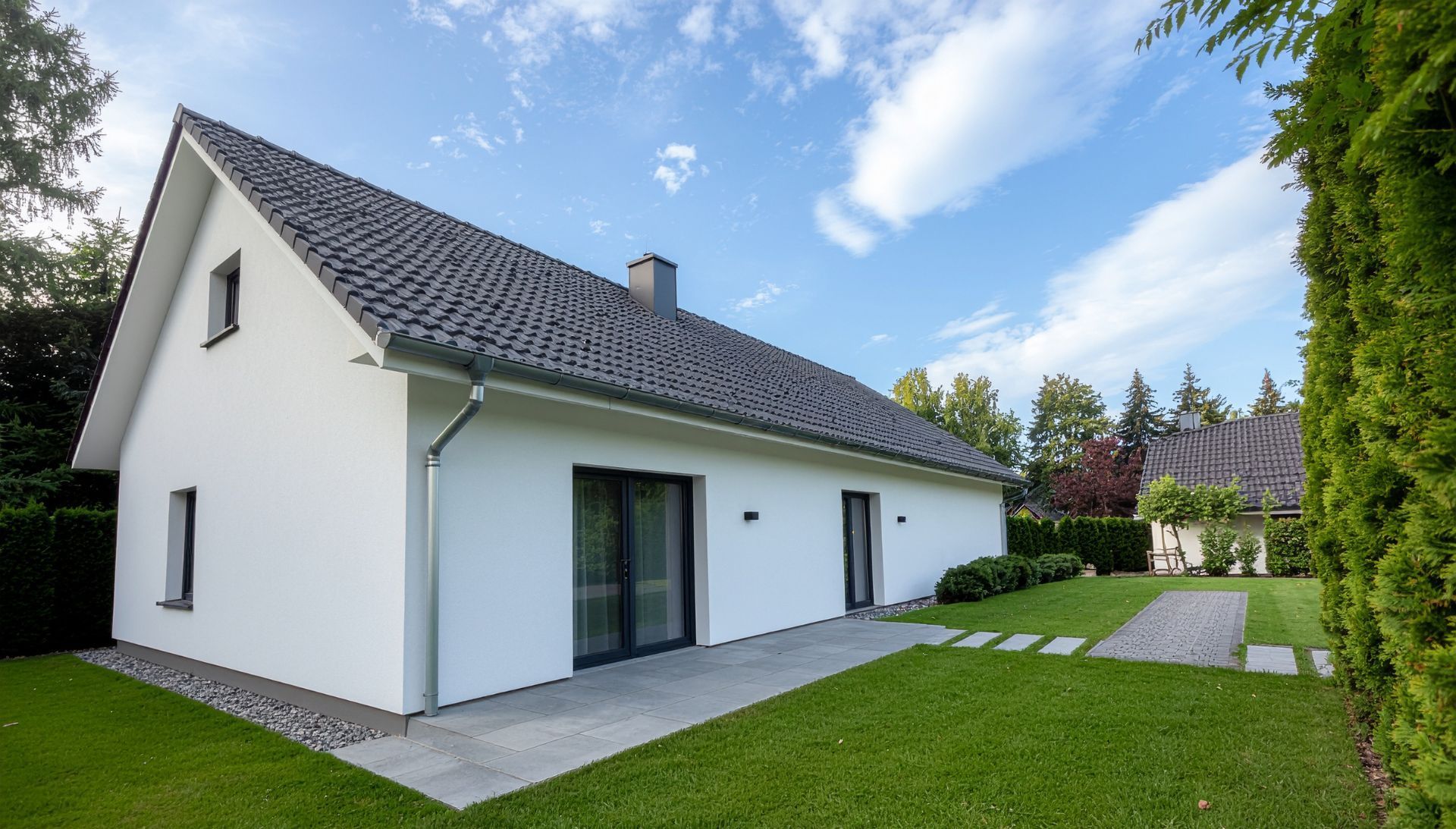 Maison blanche au toit gris, cheminée et porte-fenêtre coulissante, située sur une pelouse verte sous un ciel bleu.