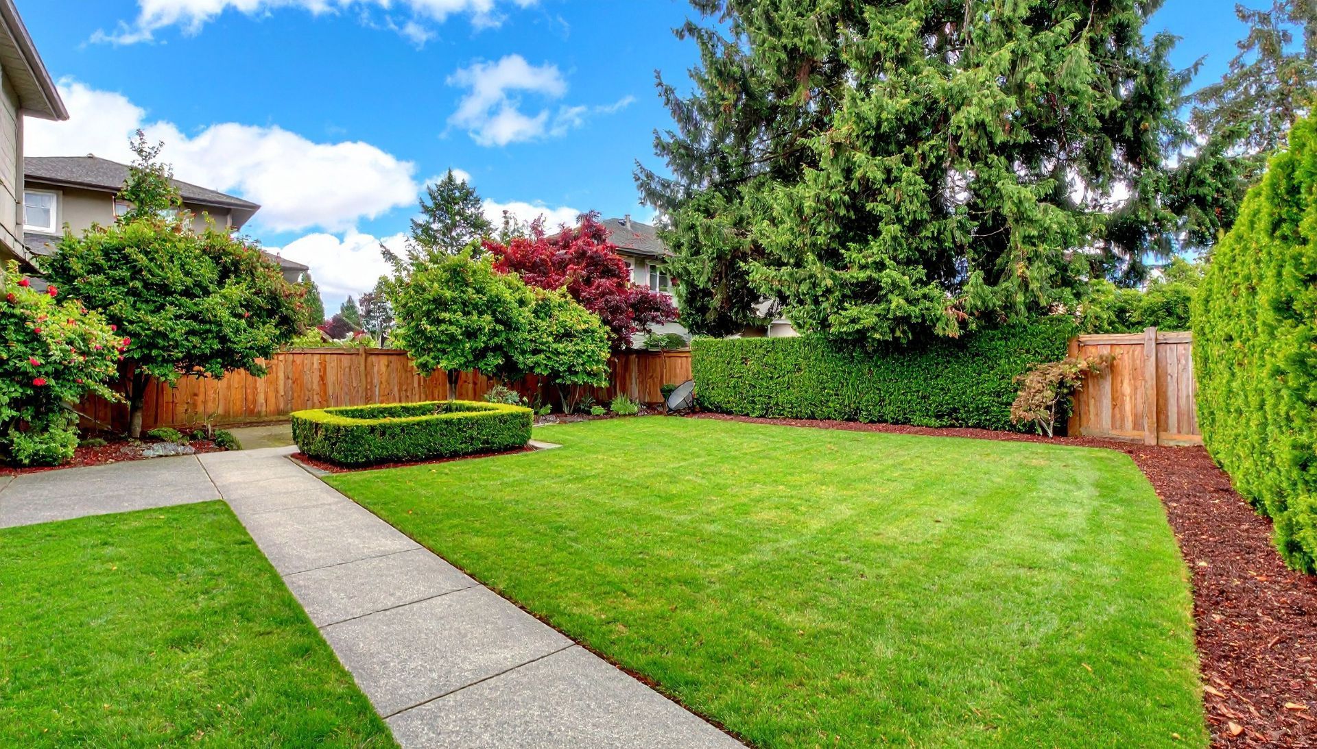 Un jardin arrière bien entretenu avec une pelouse verte, un chemin en béton, une clôture en bois.