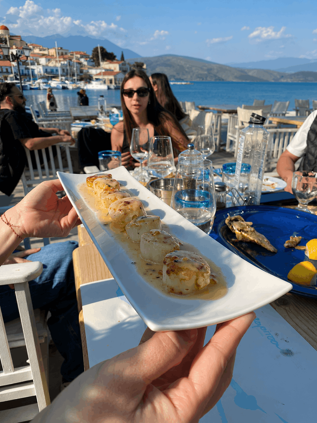 Χέρι που κρατά ένα πιάτο με επιδόρπια σε μια παραθαλάσσια καφετέρια με ποτά και ηλιόλουστη θέα στο λιμάνι
