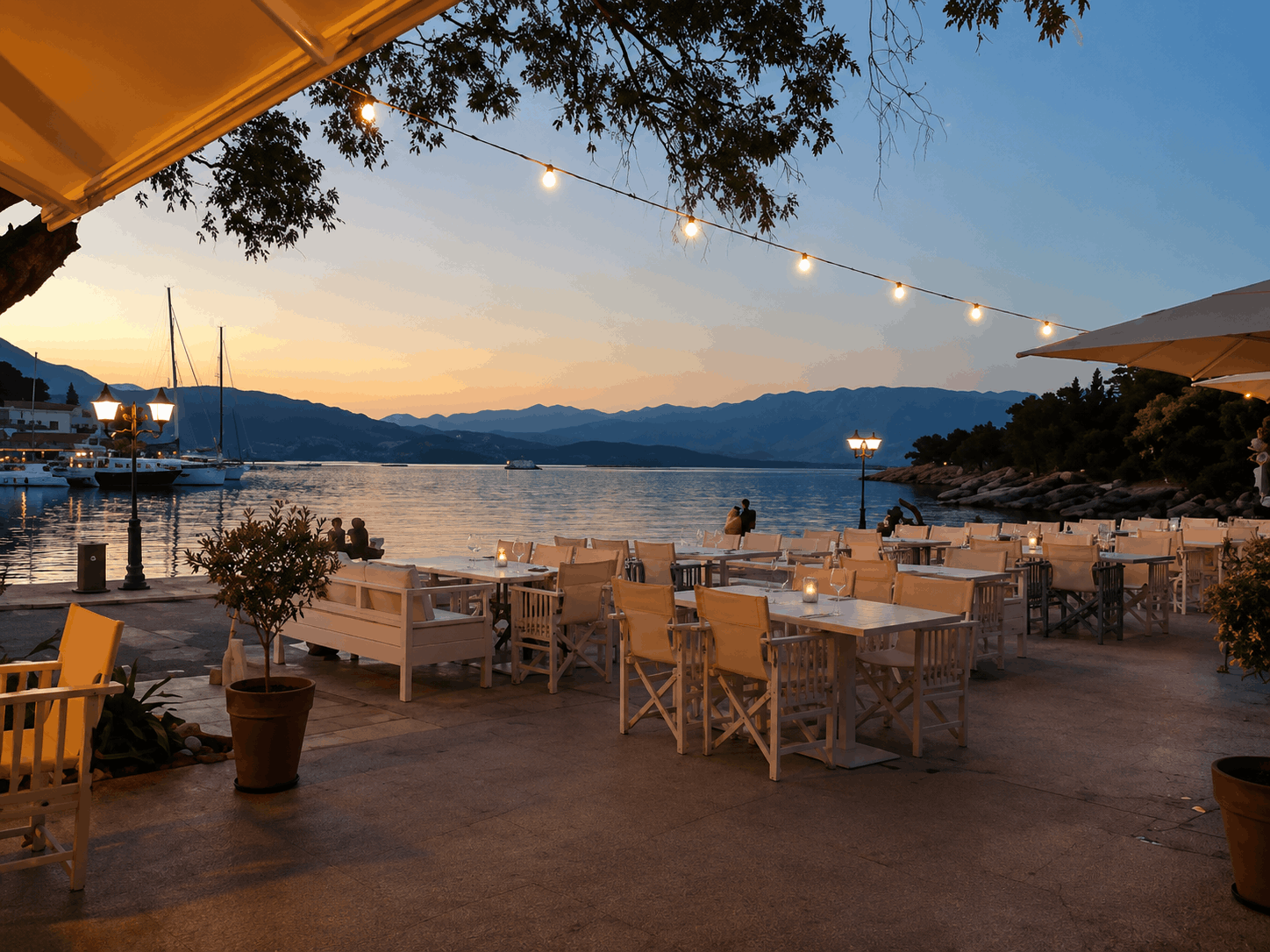 Παραθαλάσσια βεράντα εστιατορίου το σούρουπο με φωτάκια από string, λευκά τραπέζια και καρέκλες, και ήρεμα νερά από πίσω.