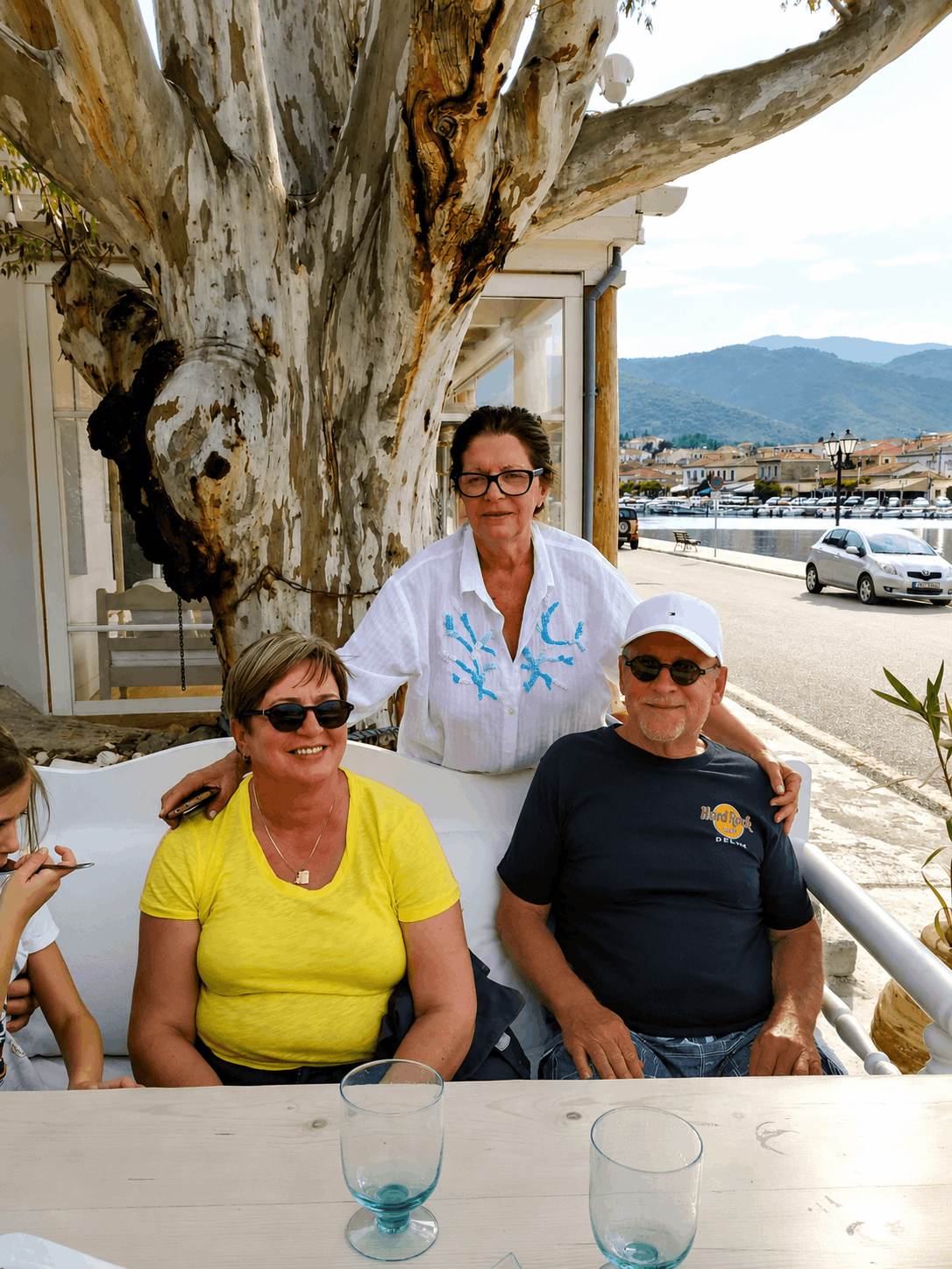 Τρία άτομα κάθονται σε ένα τραπέζι εξωτερικού χώρου καφέ δίπλα στη θάλασσα με έναν σερβιτόρο να στέκεται πίσω τους.