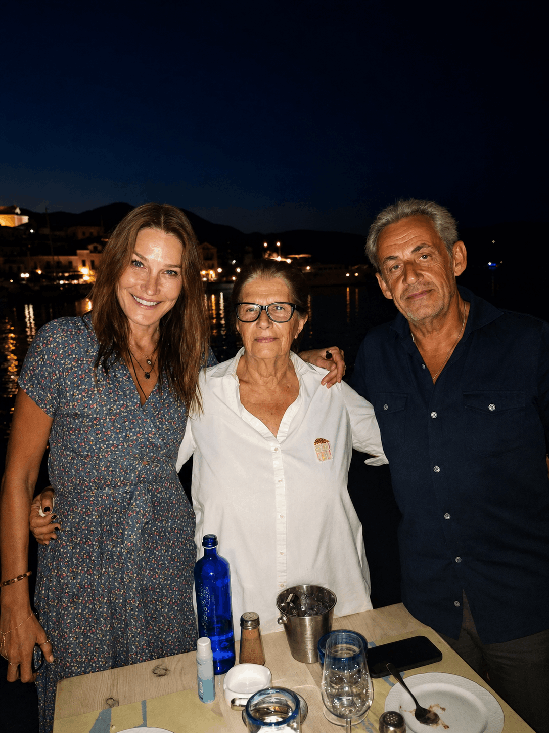 Τρία άτομα χαμογελούν σε ένα εξωτερικό τραπέζι τη νύχτα, με ποτά και ένα μπλε μπουκάλι.
