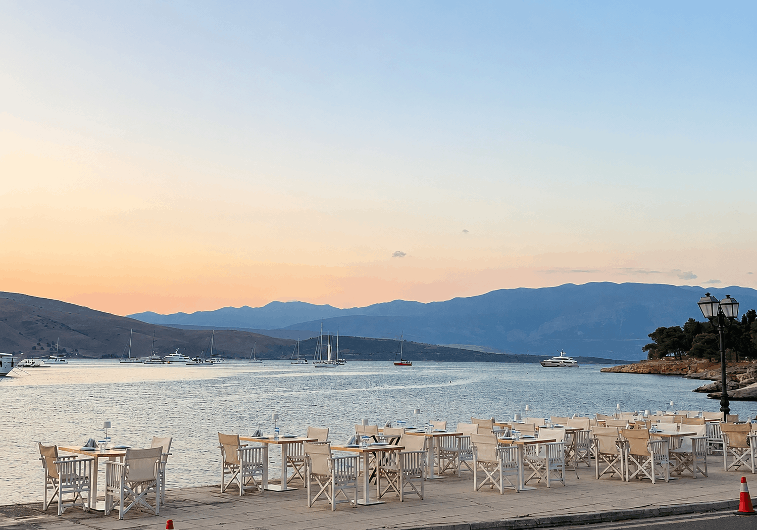 Τραπεζάκια σε καφέ μπροστά στην παραλία με θέα σε έναν ήρεμο κόλπο κατά το ηλιοβασίλεμα, με τα βουνά στο βάθος.Εστιατόριο Μαρίτσα