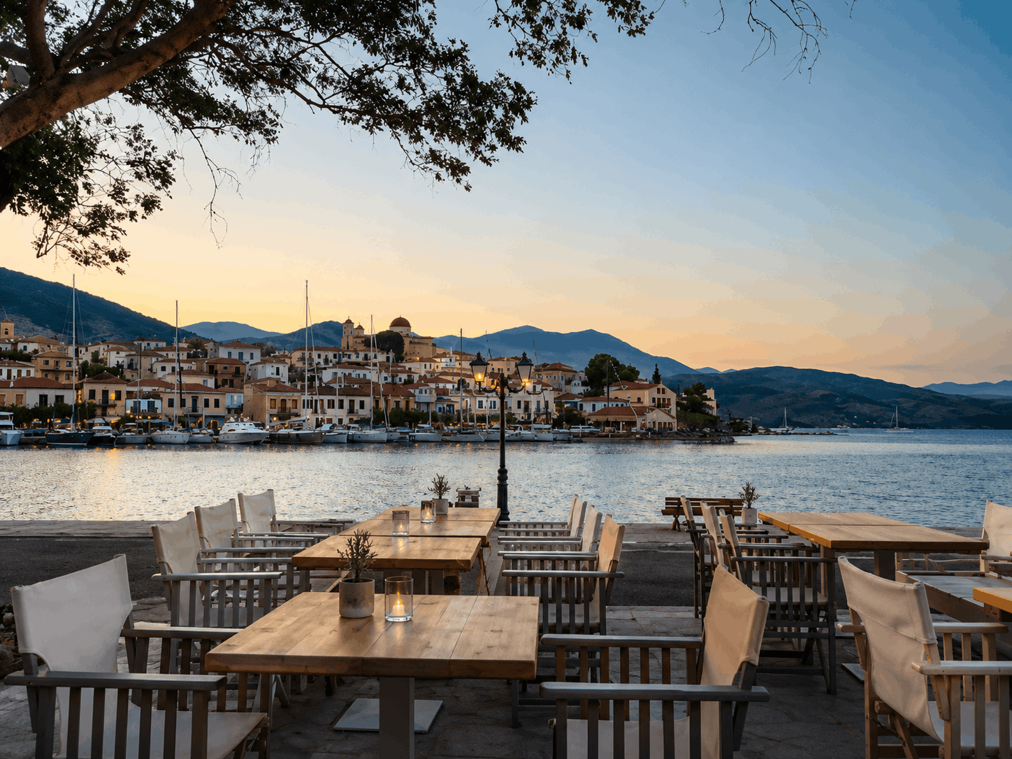 Τραπέζια καφέ δίπλα στη θάλασσα με θέα σε ένα ήρεμο λιμάνι κατά το ηλιοβασίλεμα, με βουνά και βάρκες στο βάθος