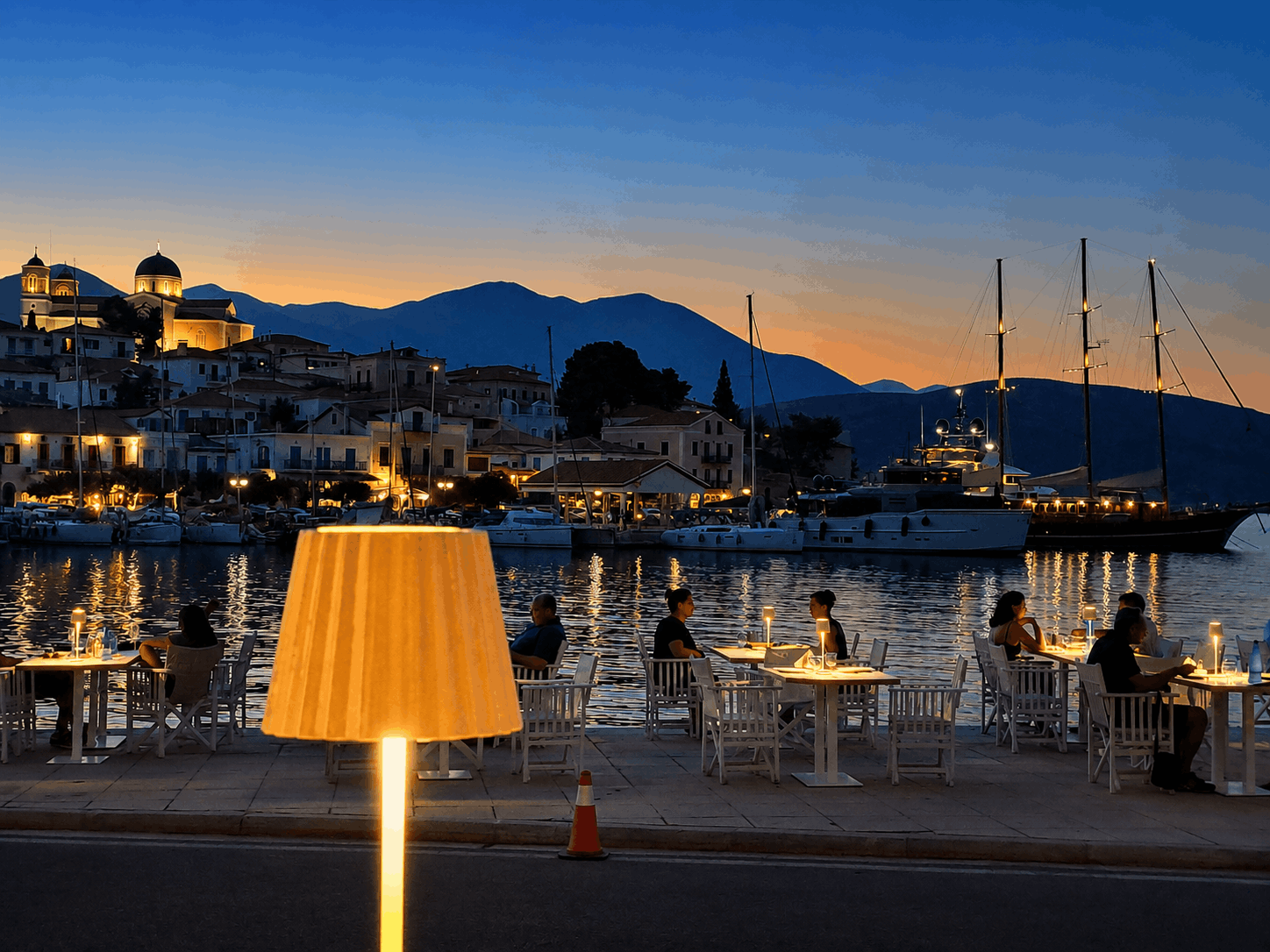 Βραδινό παραθαλάσσιο καφέ με αναμμένες επιτραπέζιες λάμπες, αγκυροβολημένο ιστιοφόρο και βουνά στο ηλιοβασίλεμα