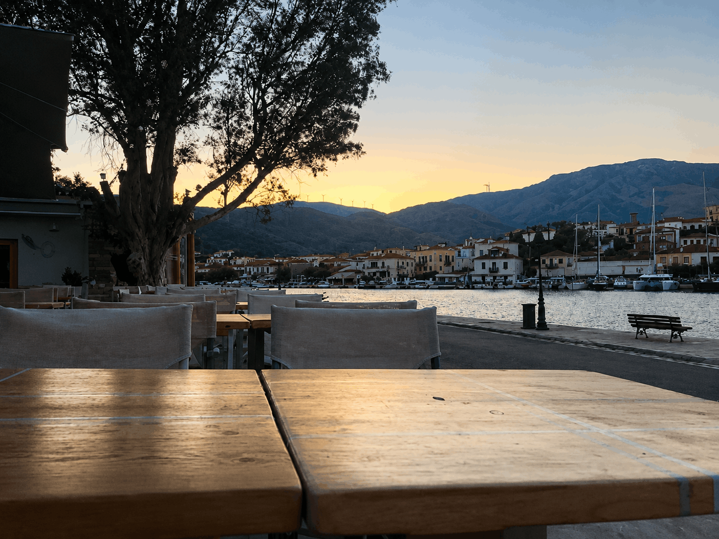 Τραπέζια καφέ δίπλα στο νερό κατά τη δύση του ηλίου με βουνά και βάρκες στο βάθος
