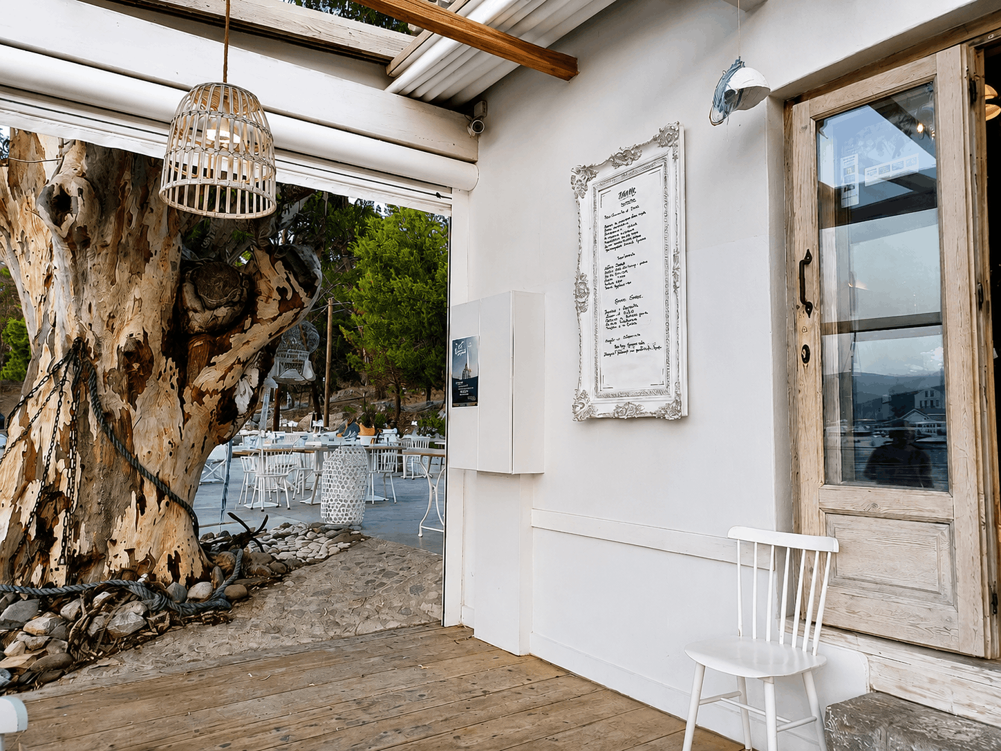 Λευκή βεράντα με ρουστίκ ξύλινη πόρτα, κρεμαστό φωτιστικό και ένα μεγάλο δέντρο ορατό από μια ανοιχτή πλευρά.