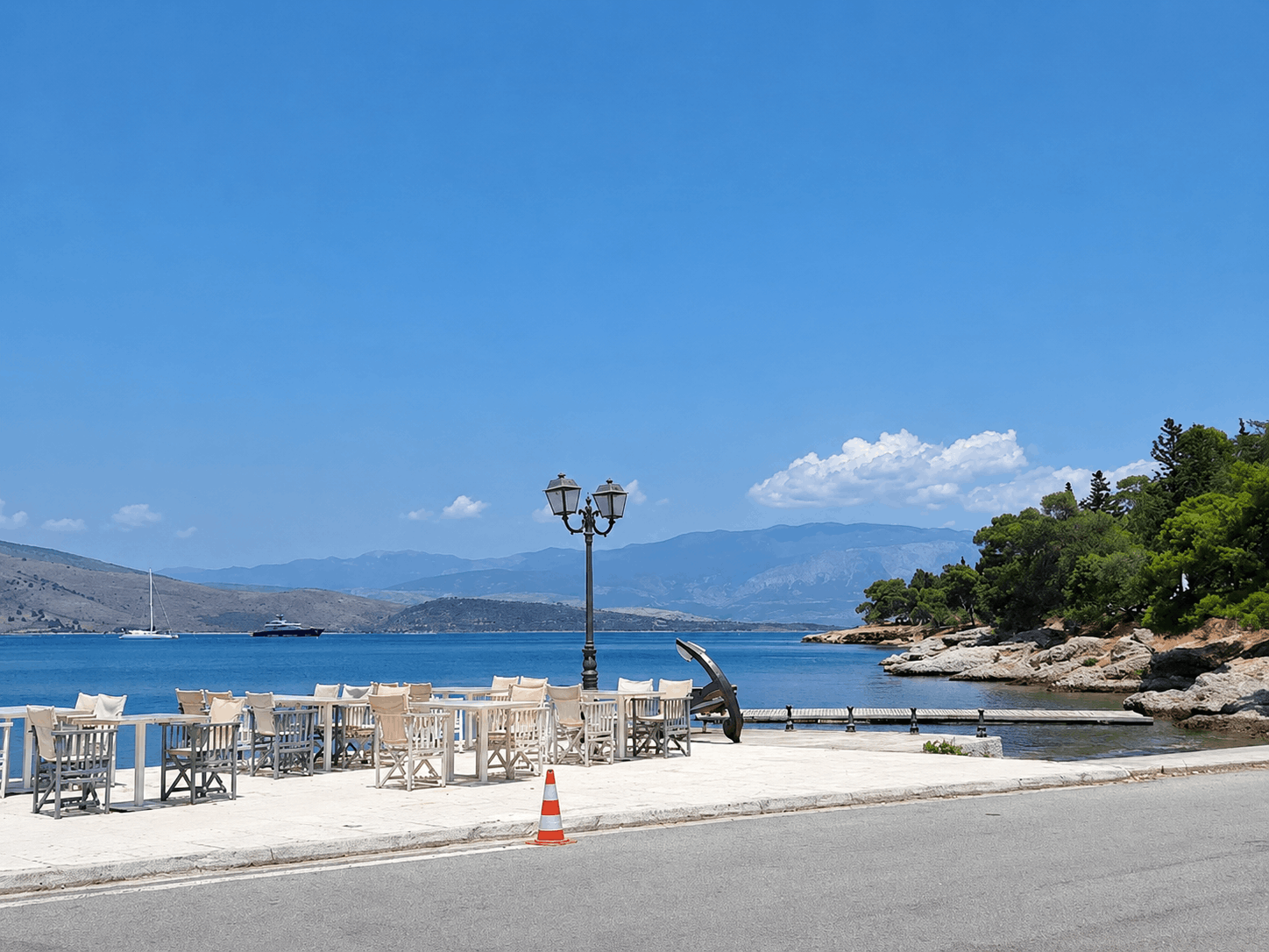 Ηλιόλουστη παραθαλάσσια βεράντα με λευκά τραπέζια, στύλο φωτισμού και γαλάζια νερά και βουνά στο φόντο