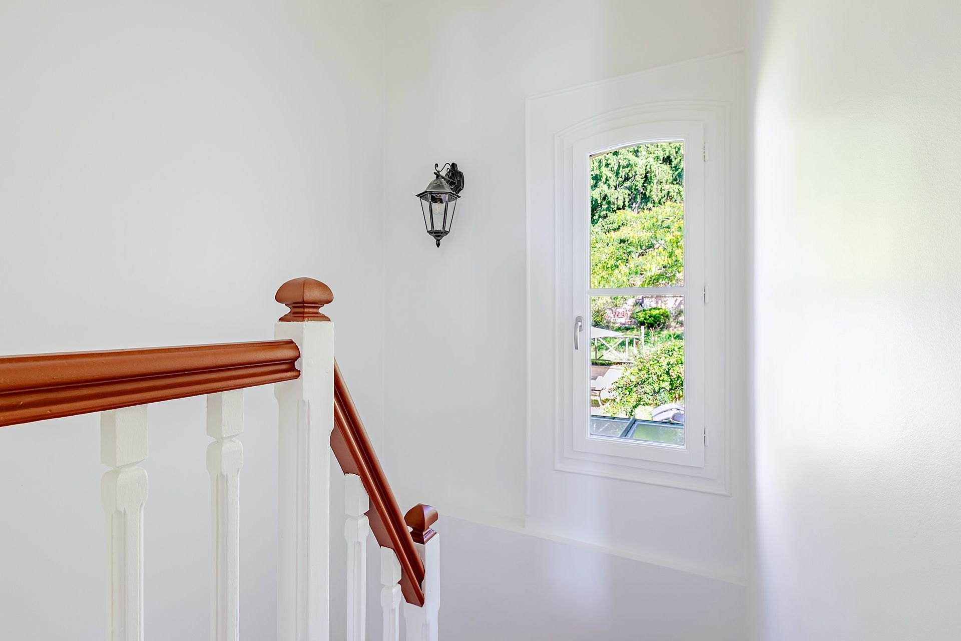 Escalier avec murs blancs, rampe en bois et fenêtre donnant sur des arbres verts.