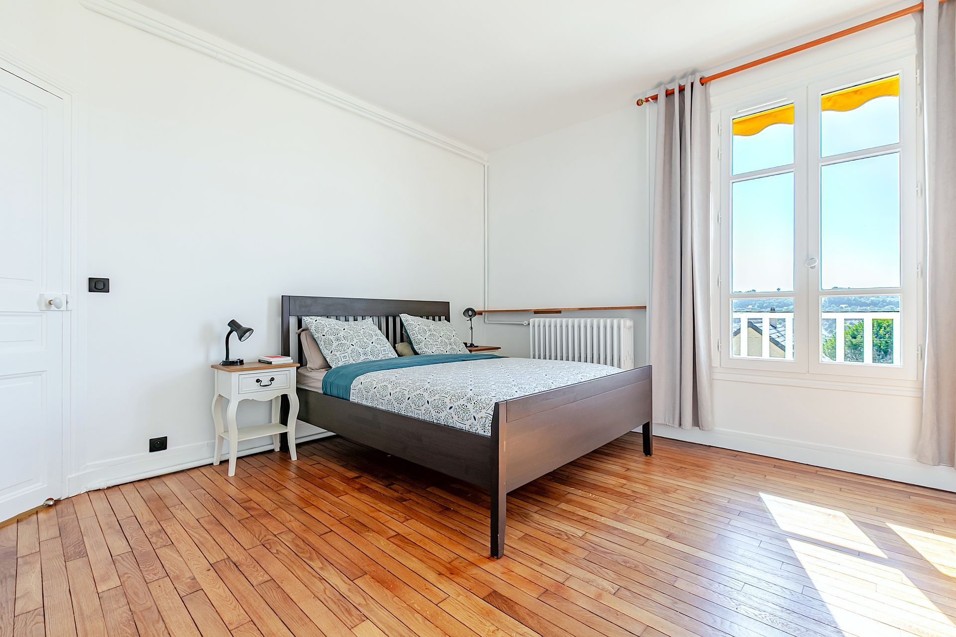 Chambre avec du parquet, un lit foncé, une petite table de chevet et une grande fenêtre avec un auvent jaune.