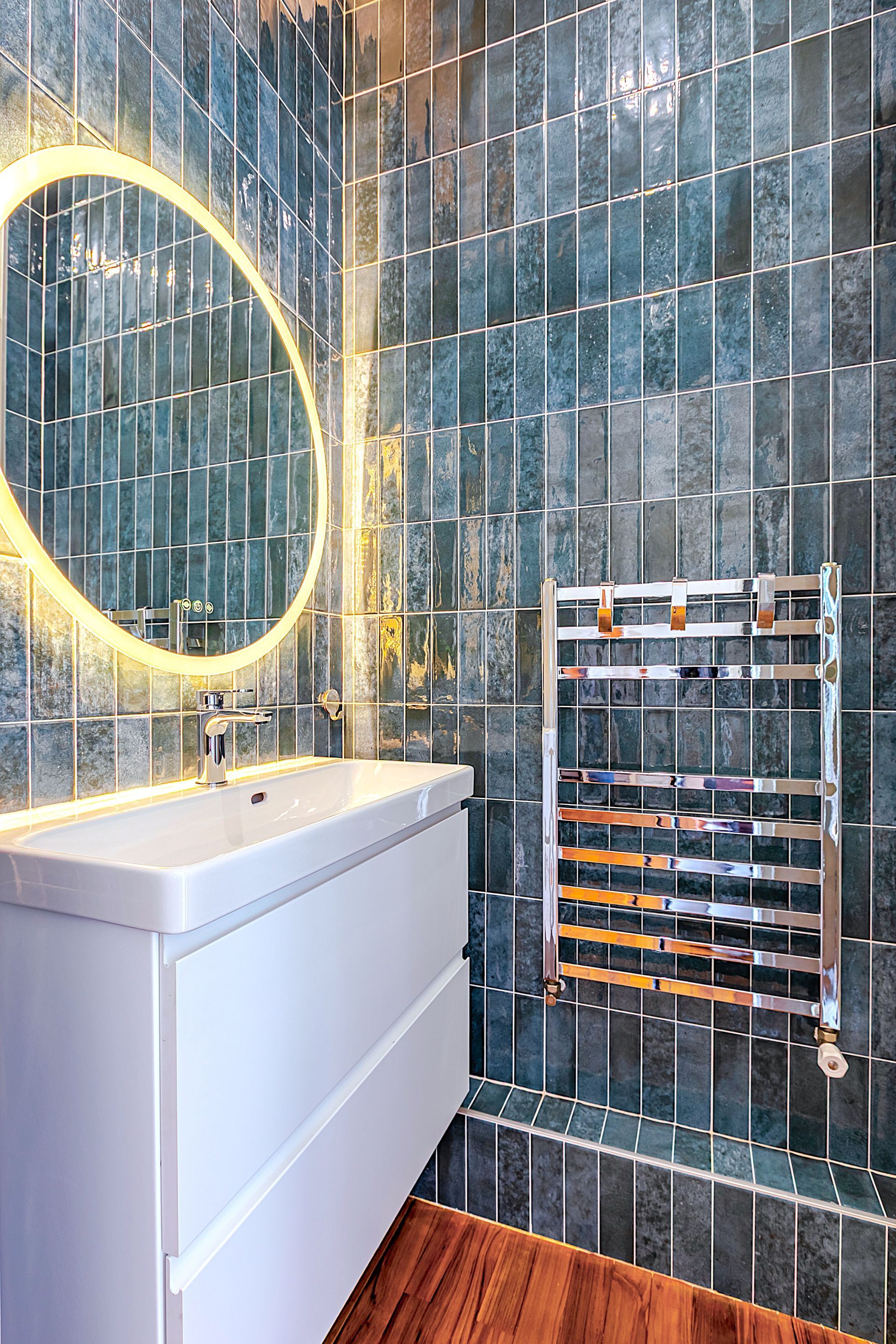 Salle de bains moderne avec mur en carrelage bleu, miroir éclairé, lavabo blanc et chauffe-serviettes chromé.