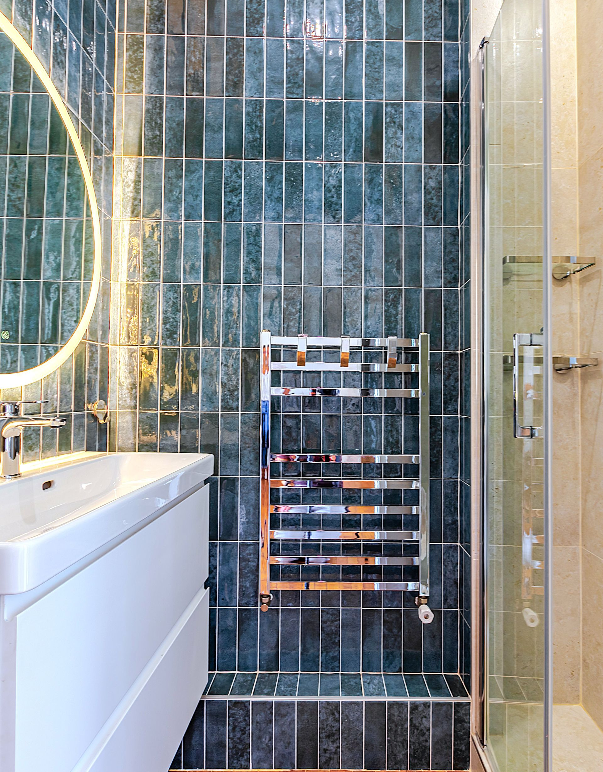 Salle de bains avec carrelage bleu sarcelle, sèche-serviettes chromé, lavabo blanc et douche.
