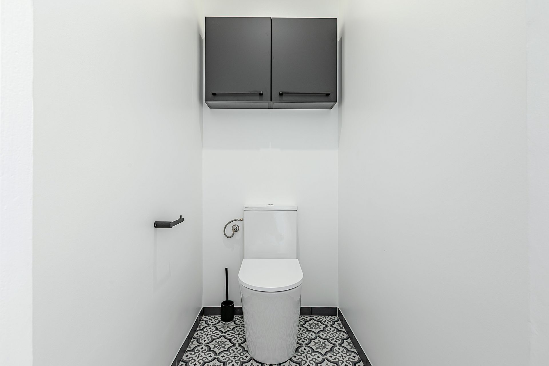 Petite salle de bains blanche avec toilettes et meuble gris foncé au-dessus. Carrelage à motifs noirs et blancs au sol.