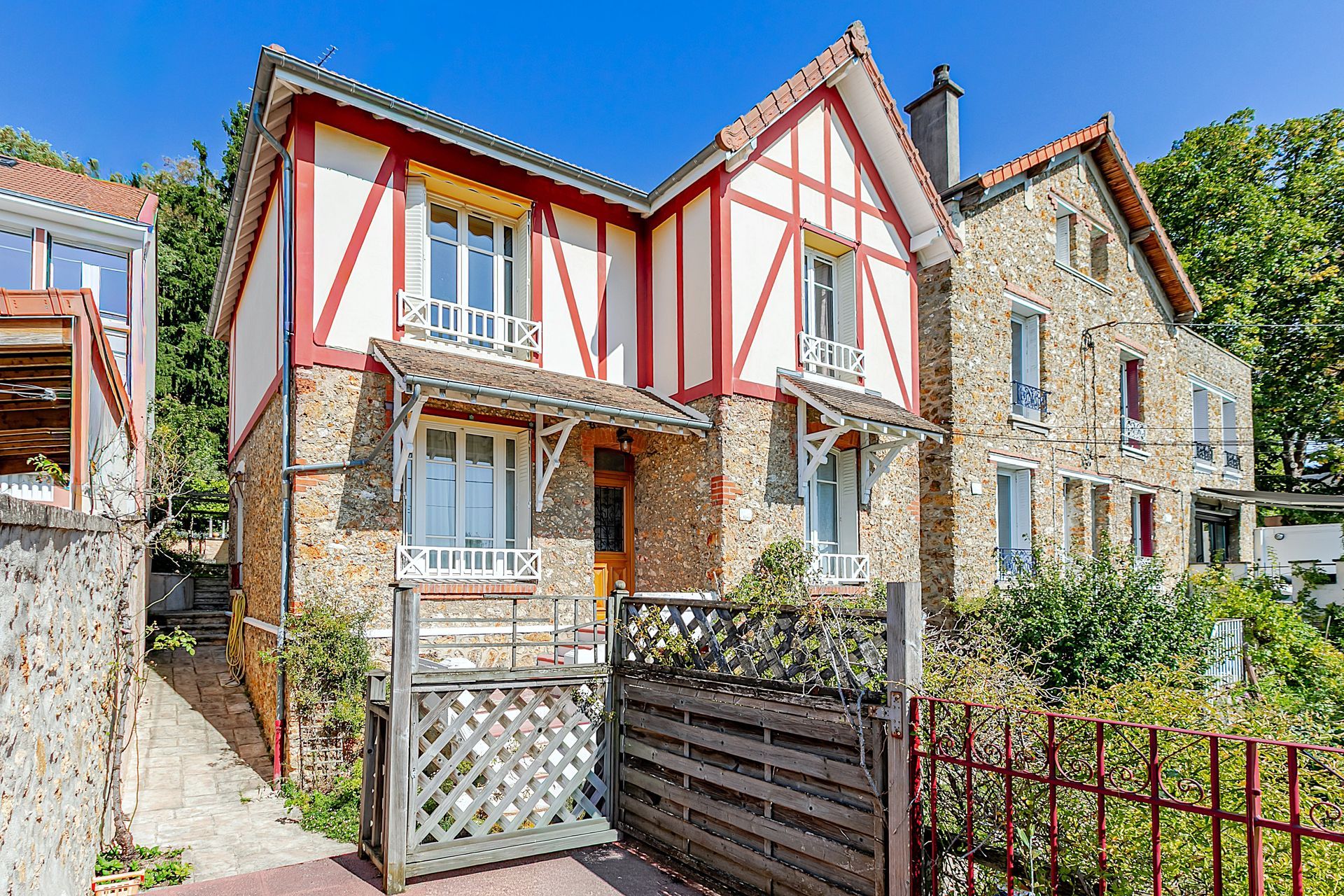 Maison à deux étages avec des colombages rouges, une façade en pierre et un brise-vue en bois.