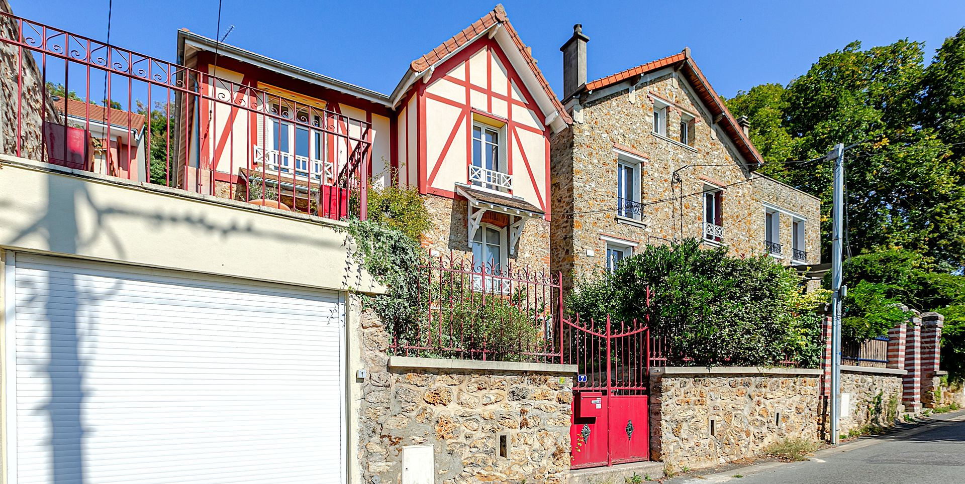 Maisons en pierre et à colombages derrière un muret en pierre, un portail rouge et une porte de garage blanche.