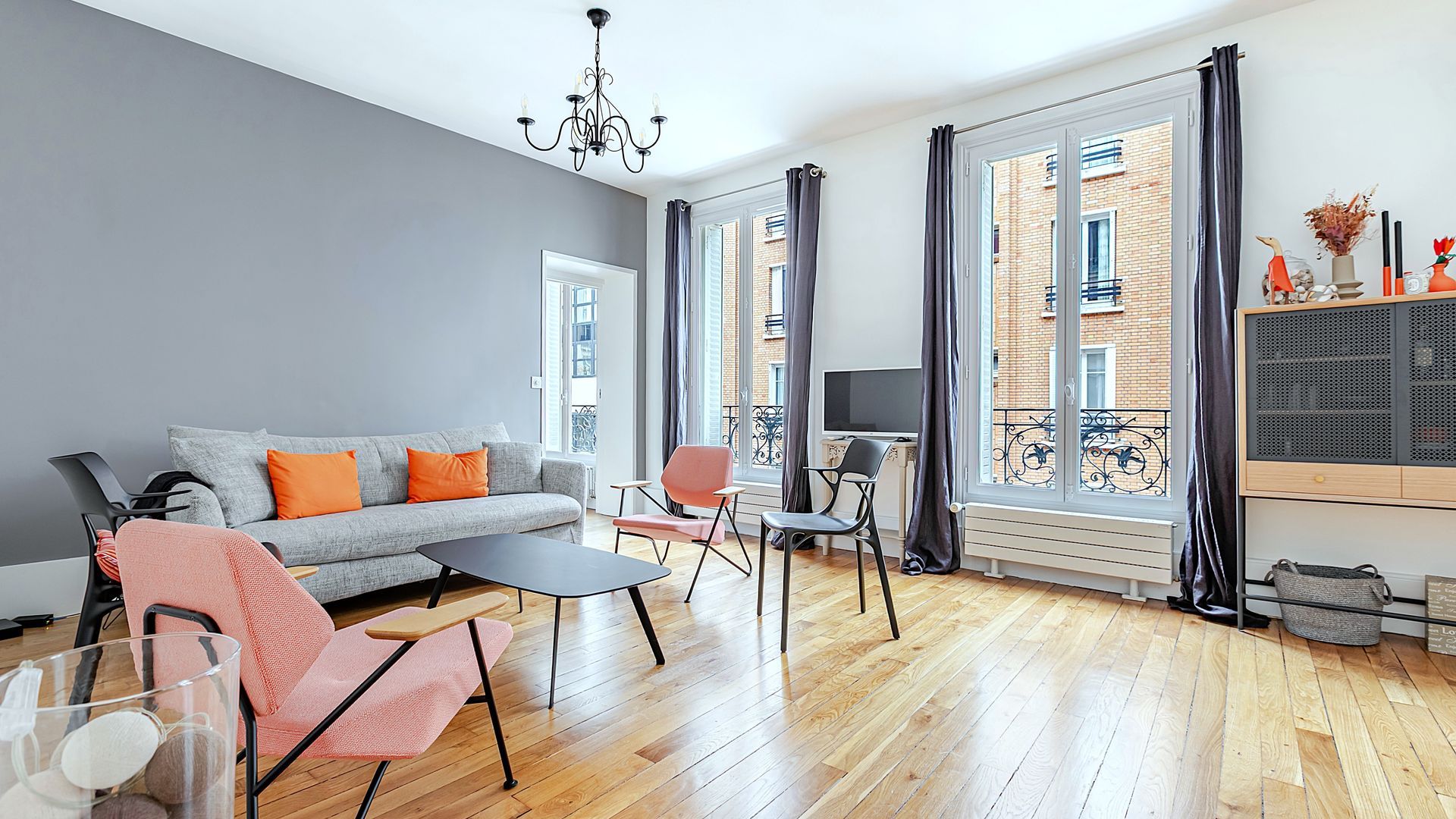 Salon avec mur gris, canapé avec coussins orange, chaises roses et grandes fenêtres.
