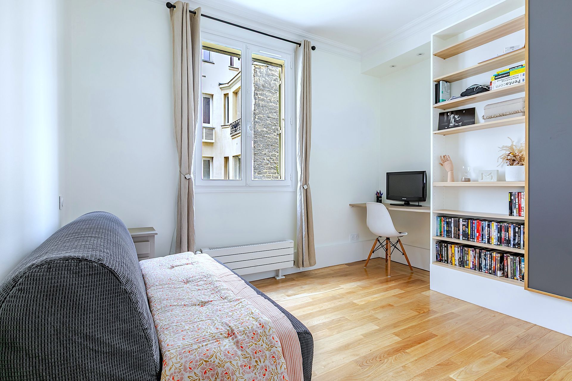 Petite pièce avec canapé, bureau, étagères et fenêtre donnant sur les bâtiments.