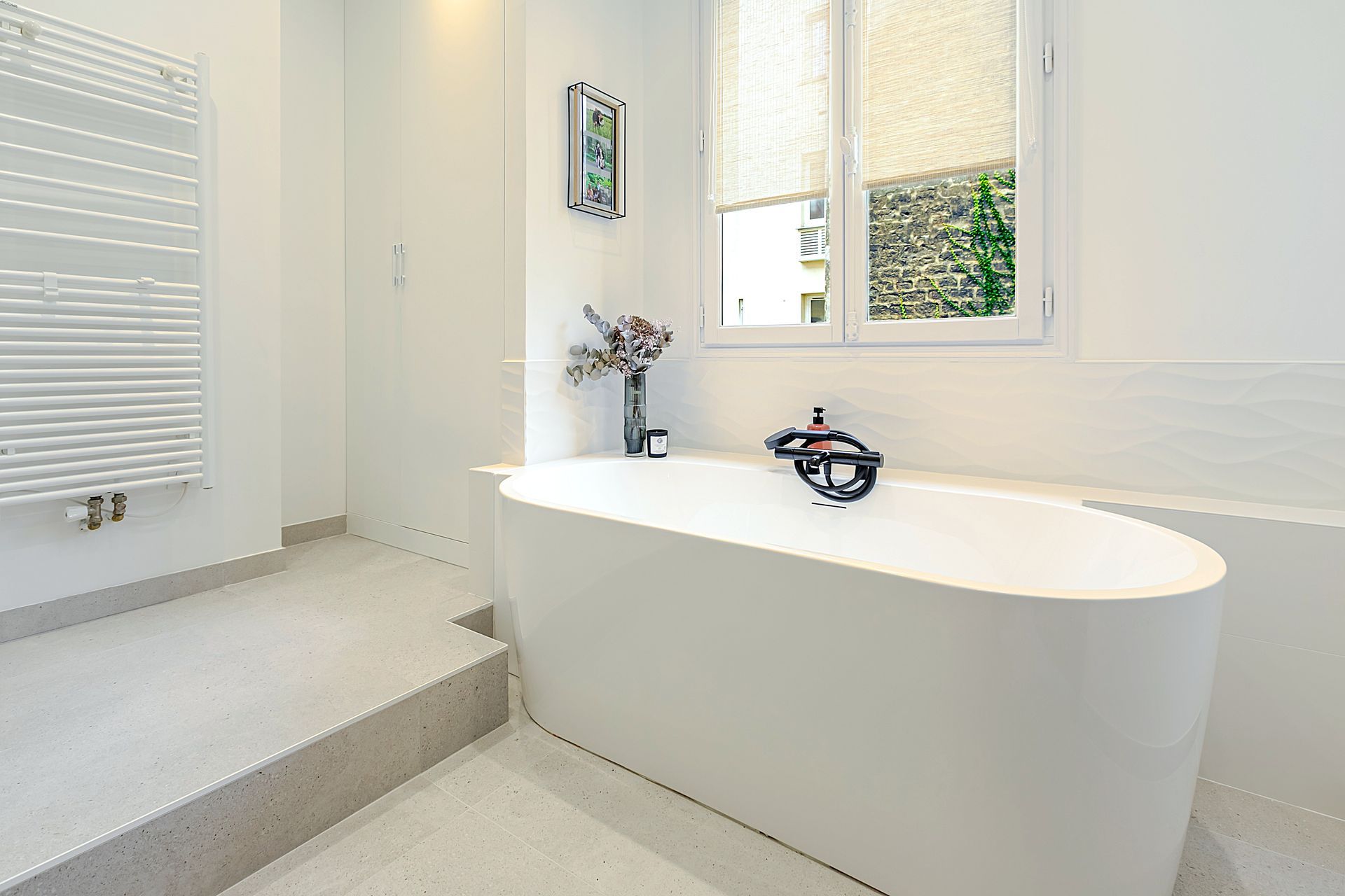 Salle de bain blanche avec baignoire ovale, sèche-serviettes et petite fenêtre avec vue sur la verdure.