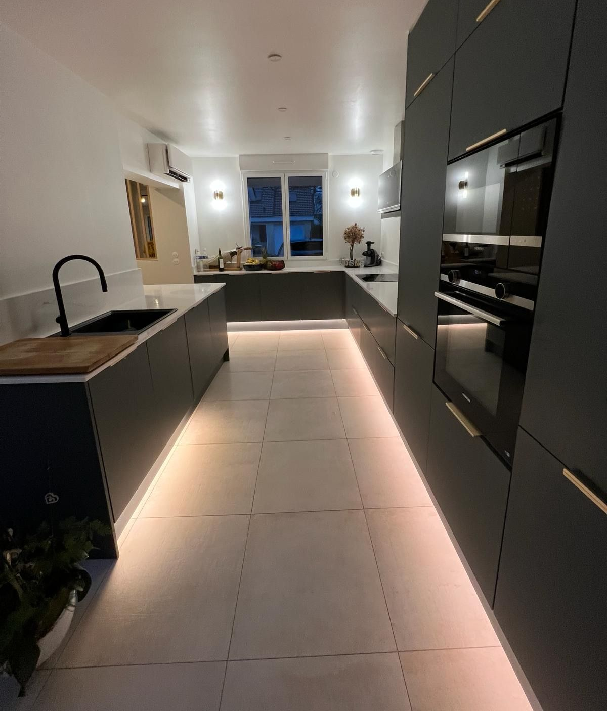 Cuisine moderne gris foncé avec éclairage sous les meubles. Évier et four noirs, poignées de placard dorées.