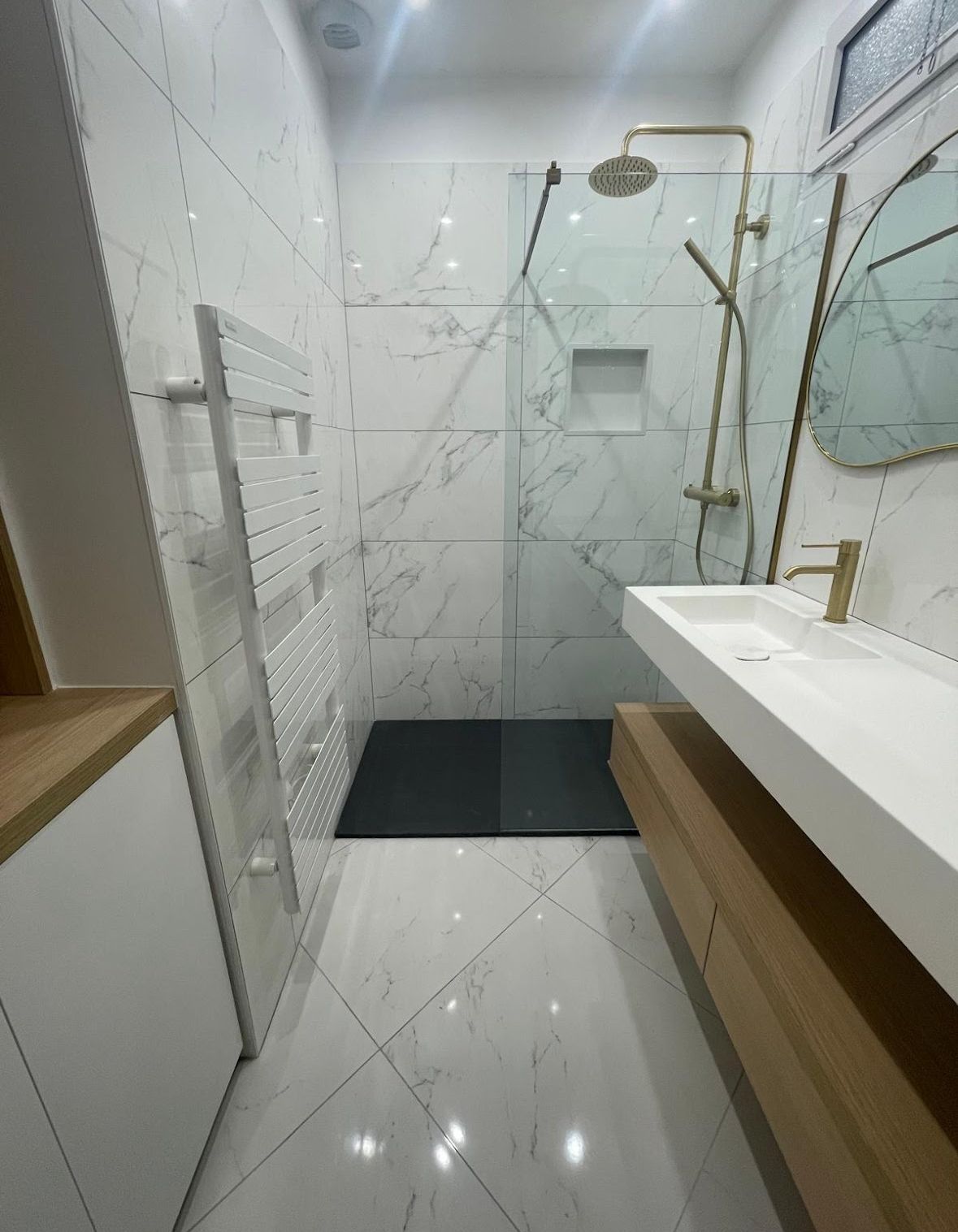 Salle de bains avec carrelage en marbre, douche en verre, luminaires dorés et vanité flottante.
