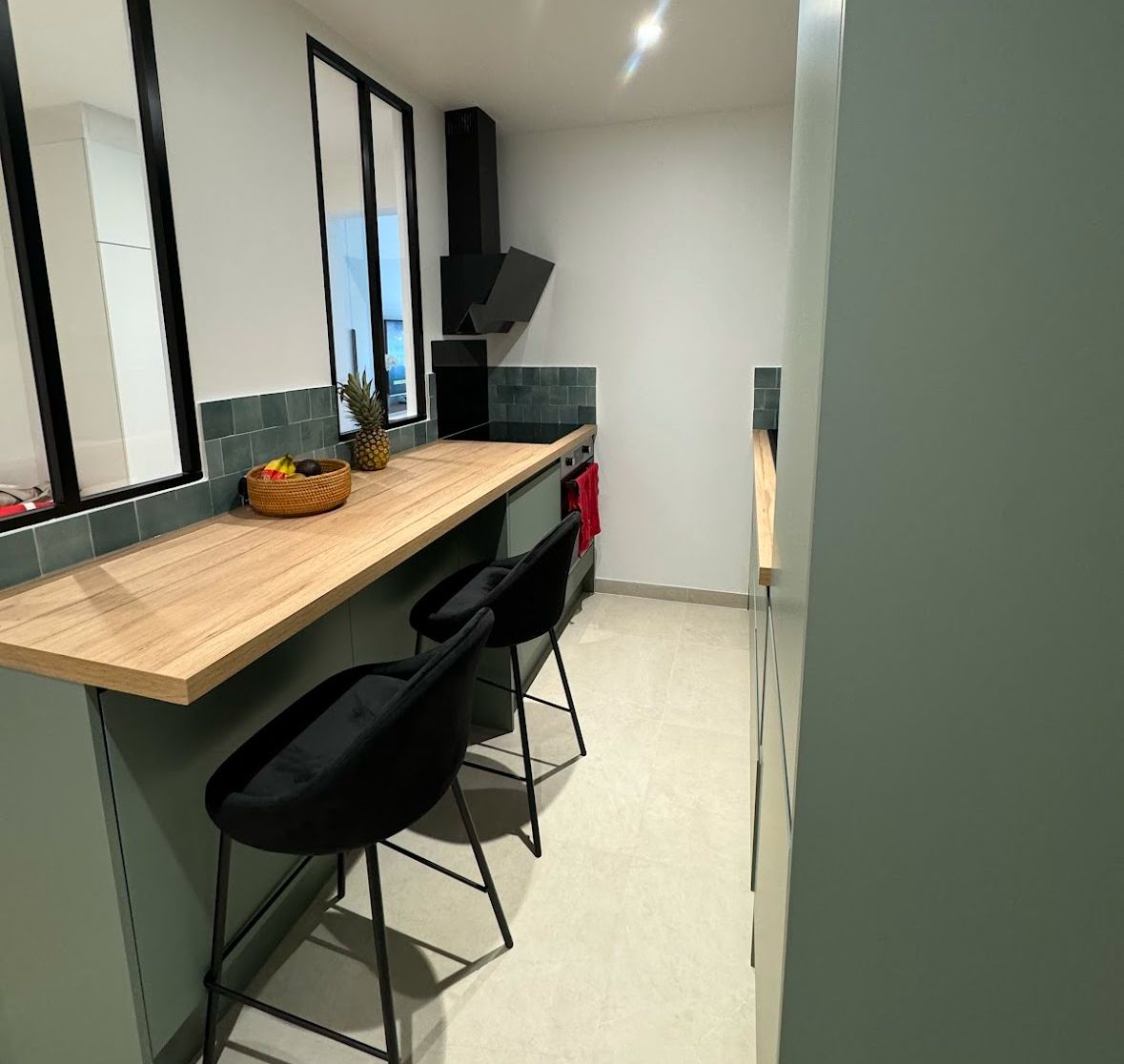 Une petite cuisine avec un comptoir en bois, deux chaises noires et une hotte noire ; une armoire vert clair avec un mur vert clair.