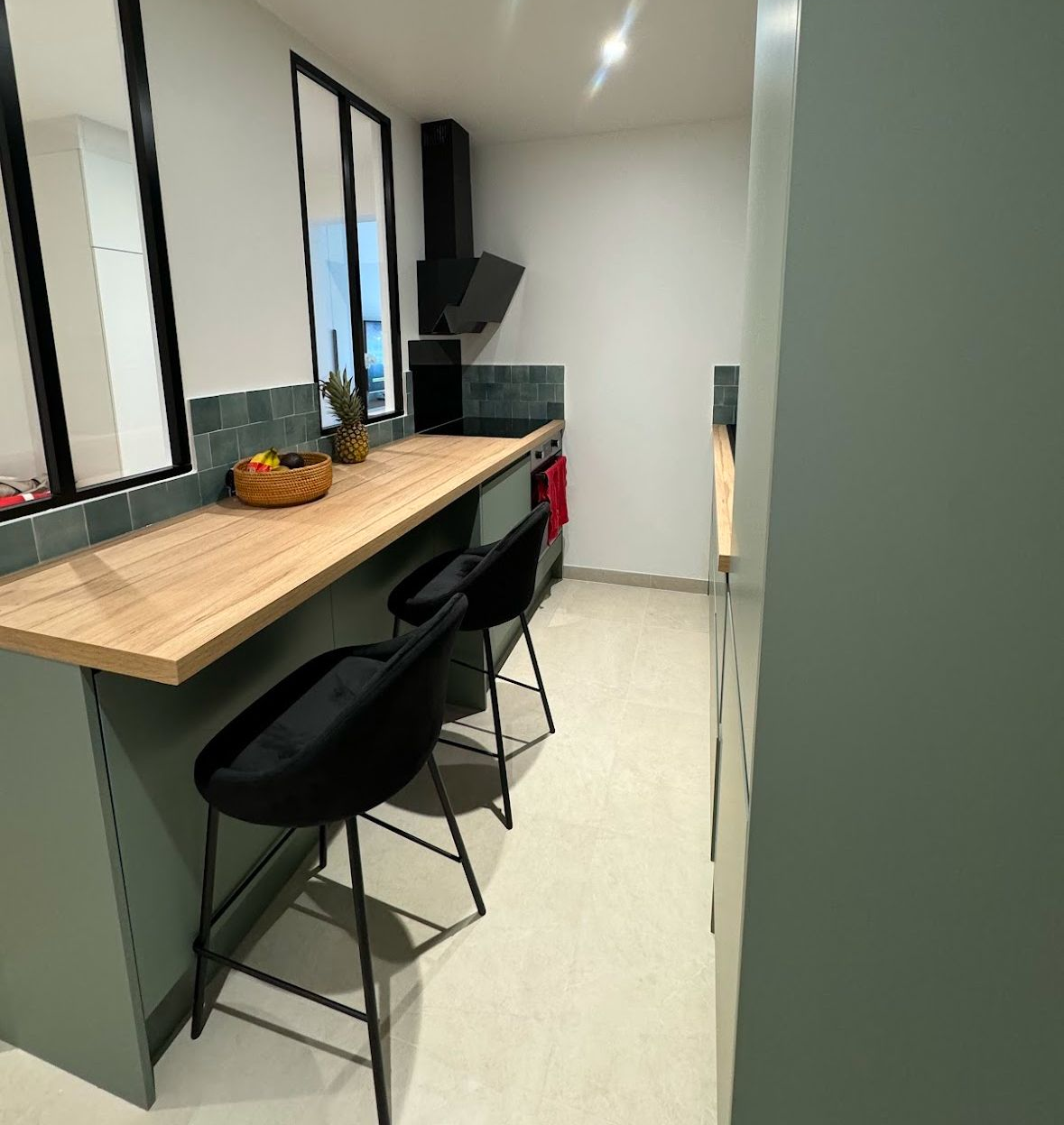 Cuisine avec armoires vertes, comptoir en bois, deux tabourets noirs.