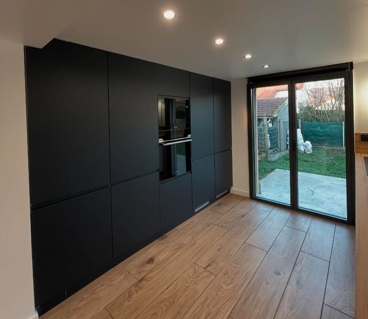 Cuisine moderne avec armoires noires, four, plancher en bois et porte vitrée donnant sur la cour arrière.